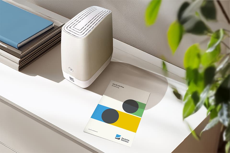 Weißer WLAN-Router auf weißem Regal neben Büchern, bunter Broschüre und grüner Zimmerpflanze.