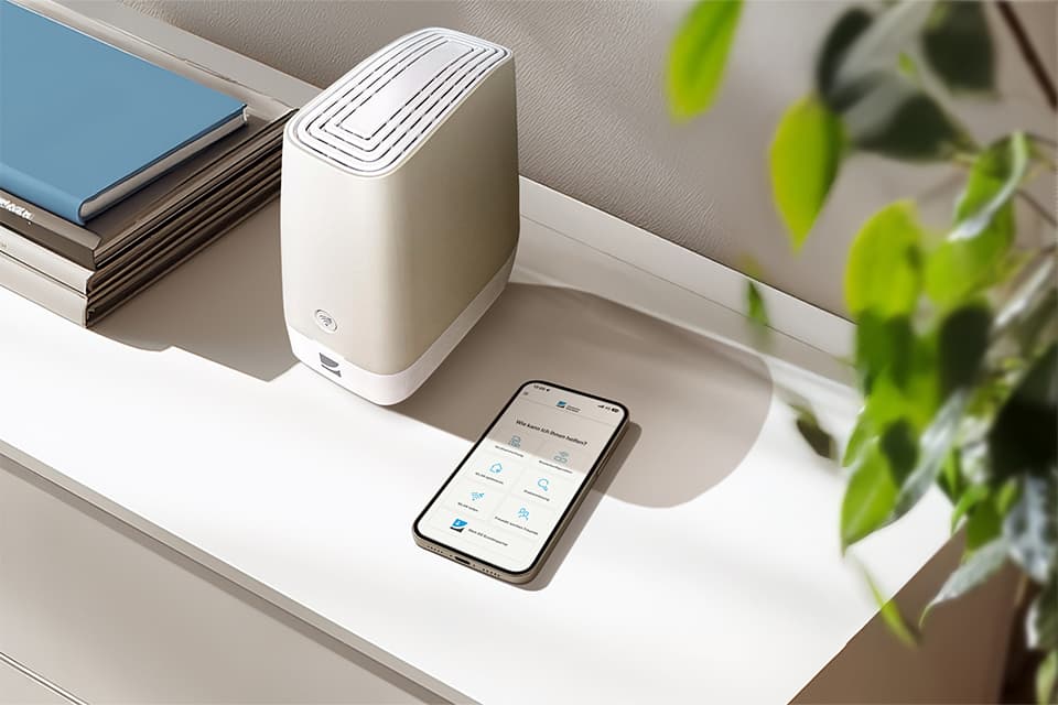 Weißer Router auf Regal neben Smartphone mit App und grüner Pflanze, blaues Buch im Hintergrund.