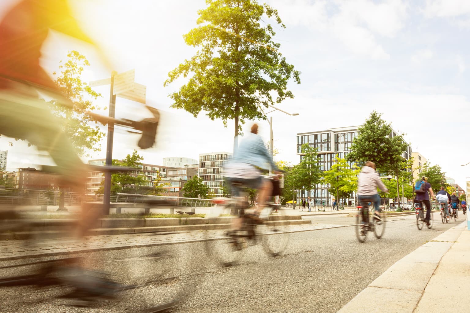Verschwommene Radfahrer auf einem Stadtradweg mit grünen Bäumen und modernen Gebäuden im Hintergrund bei sonnigem Wetter.