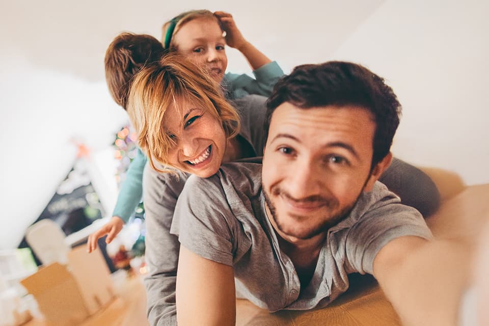 Eine Familie lächelt in die Kamera, während sie in einem hellen Wohnraum mit Umzugskartons im Hintergrund zusammen spielen.