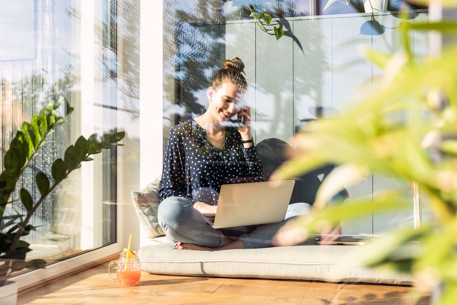 Lächelnde Frau sitzt im Schneidersitz auf Terrasse, telefoniert und arbeitet am Laptop, umgeben von Pflanzen.