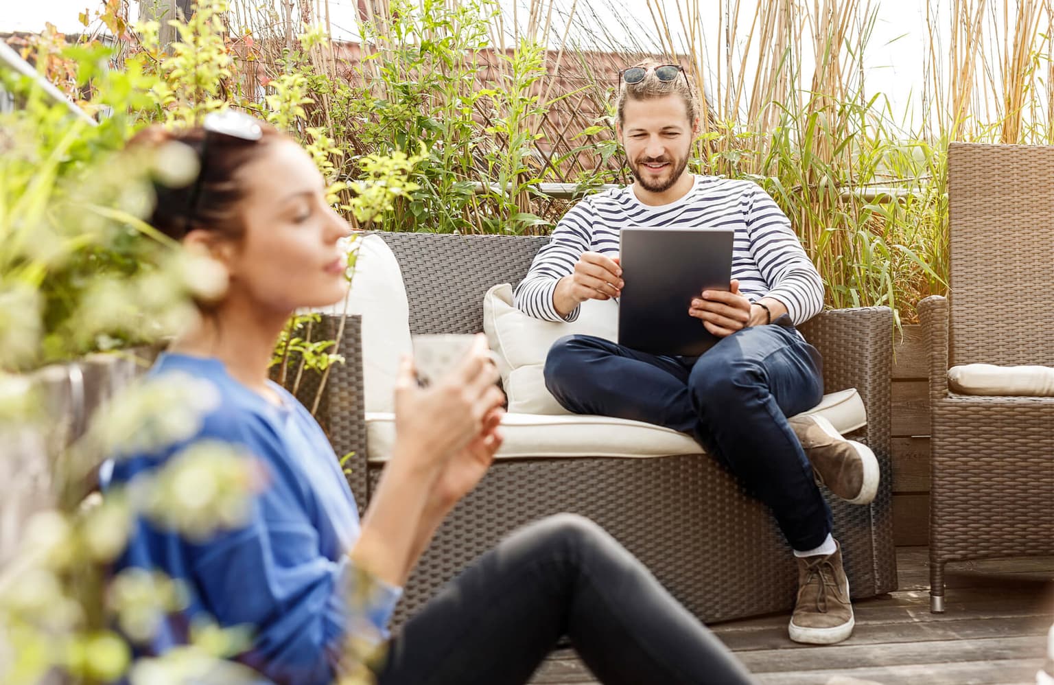 Mann und Frau sitzen bei warmen Wetter in der Sonne auf der Terrasse, während der Mann mit dem Tablet im Internet surft