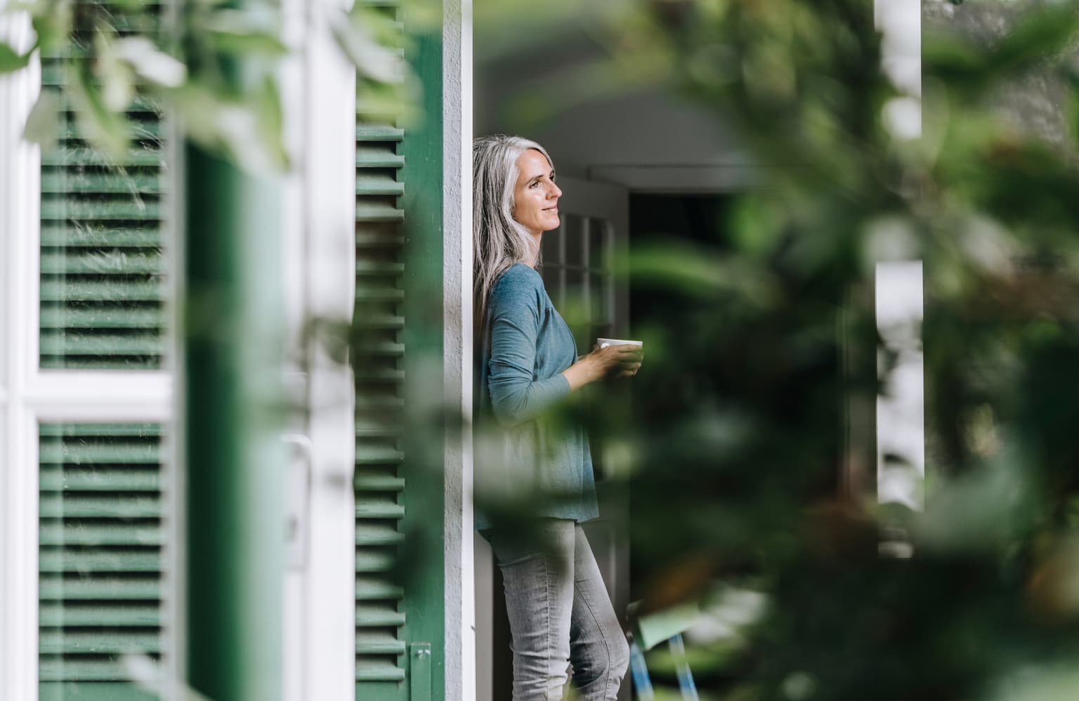 Ältere Frau steht mit Tasse nachdenklich am Fenster