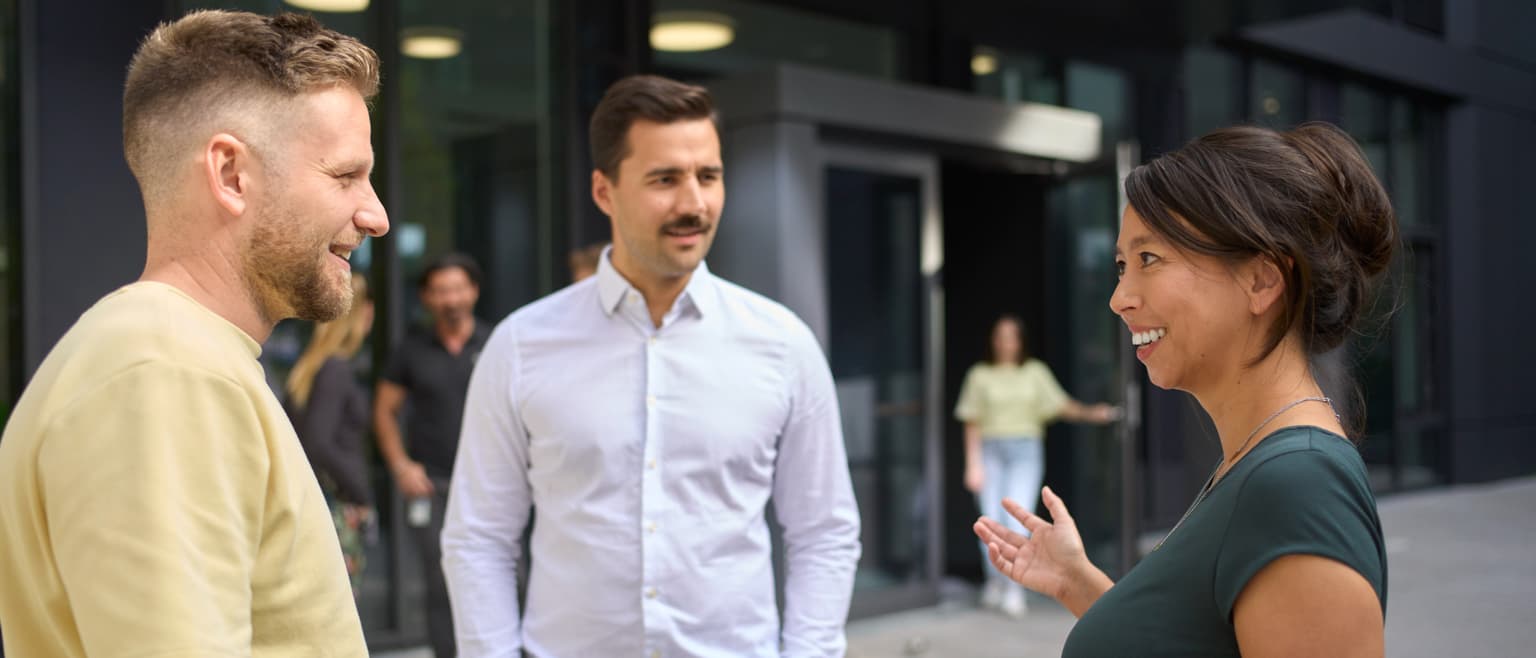 Das Bild zeigt 3 Mitarbeitende in einem Gespräch. Rechts im Bild ist eine Frau zu sehen, mittig und links jeweils ein Mann. Sie stehen vor dem Bürogebäude. Der Hintergrund ist verschwommen und dort sind noch andere Personen zu sehen, auch verschwommen.