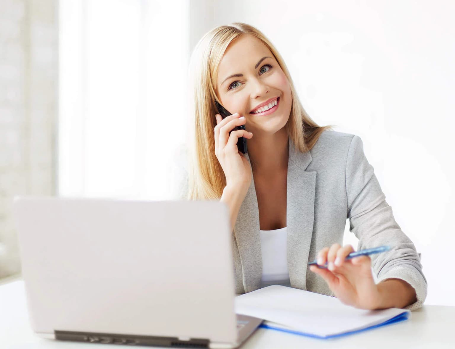 Frau im Blazer sitzt am Tisch mit Stift, Block und Laptop und telefoniert dabei mit dem Handy
