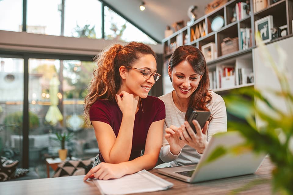 Zwei Freundinnen sitzen am laptop und schauen sich etwas auf dem Handy an