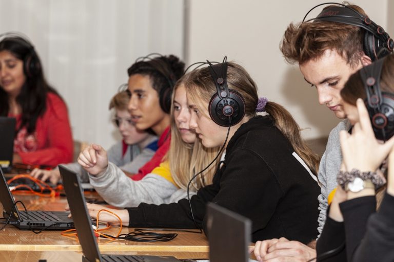 Jugendliche sitzen nebeneinander vor Laptops und tragen Kopfhörer.