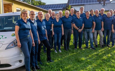 Eine Gruppe von blau gekleideten Menschen steht auf dem Rasen vor einem E-Auto mit der Aufschrift: „Wir verbinden Boxberg e. V.“