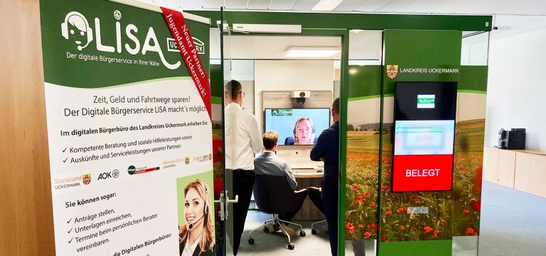 Ein gläserner Kubus mit grünen Elementen enthält Geräte für eine Live-Video-Schalte zum Bürgerbüro Uckermark: Zu sehen ist ein Monitor, vor dem drei Personen sitzen oder stehen. Im Vordergrund steht ein Informationsplakat zum Konzept LISA.