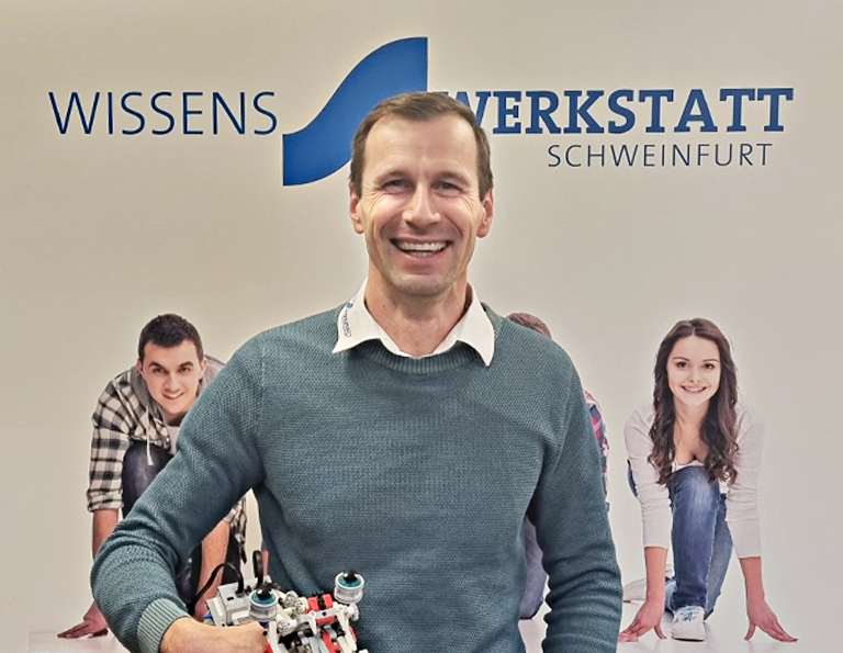 Markus Dietz steht vor einem Plakat der Wissenswerkstatt Schweinfurt und hält einen Lego-Roboter unterm Arm.