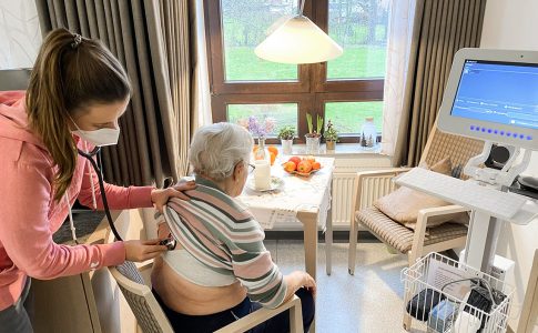 Eine alte Frau sitzt auf einem Stuhl, neben ihr ein Monitor. Eine Pflegekraft hält ein Stethoskop an den Rücken der Frau.